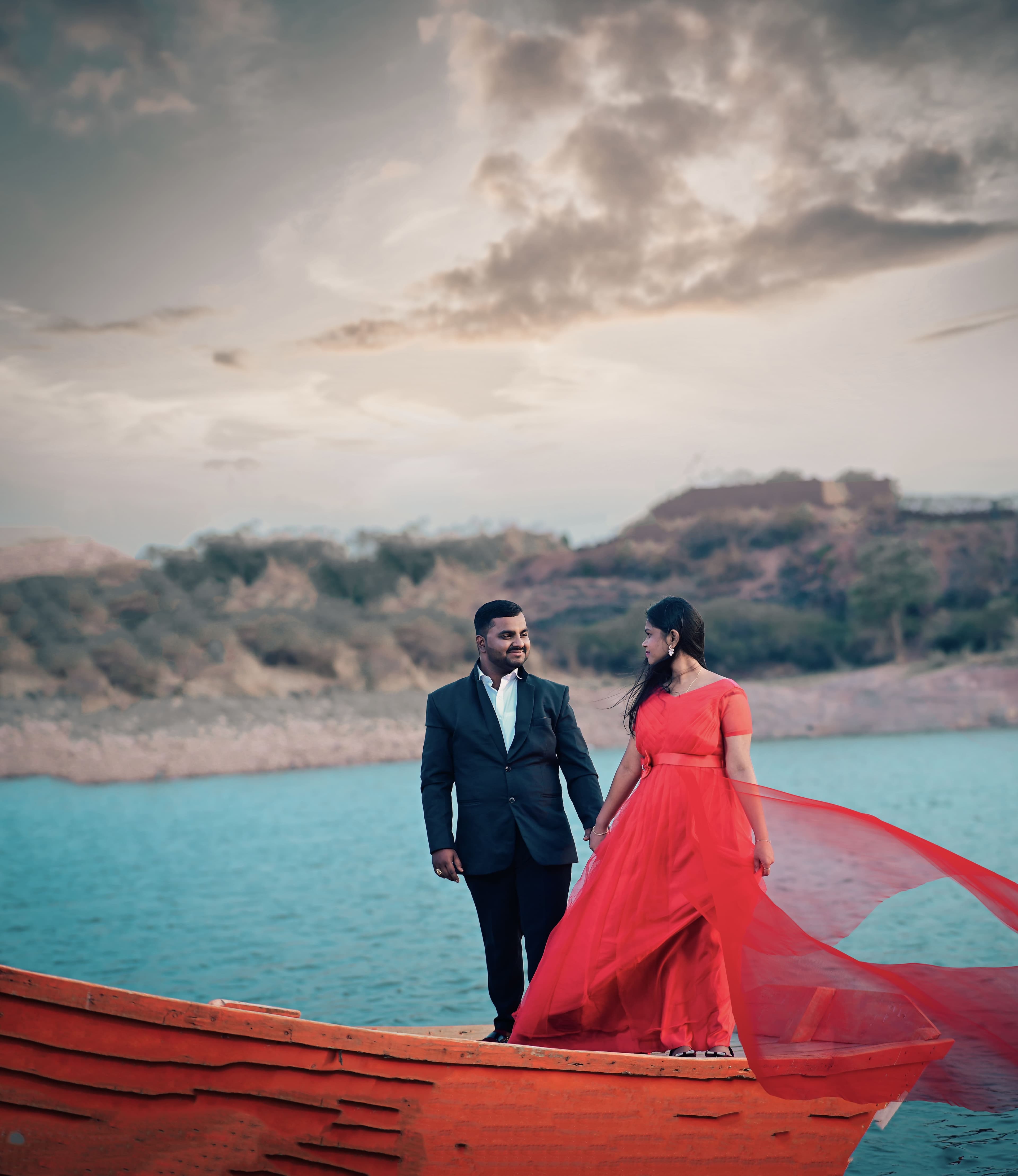 Couple on a wooden boat by a lake, red gown flowing in the wind